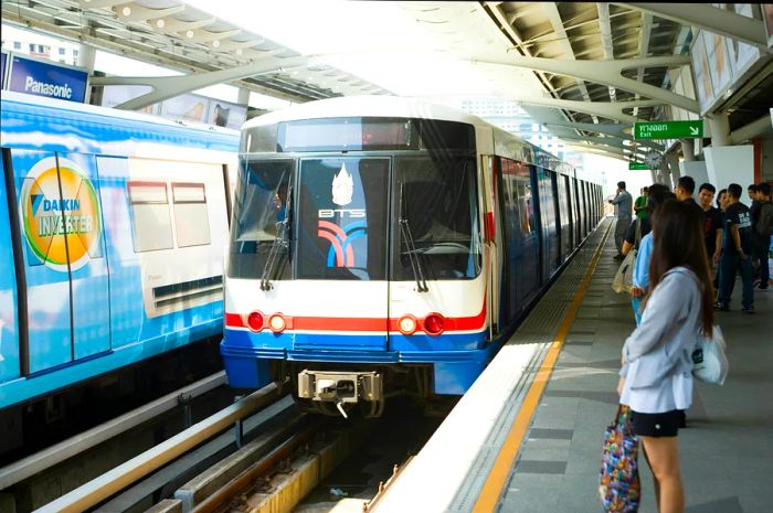 The blue, white, and red BTS Skytrain arrives at Ari station in Bangkok, as passengers move toward it to board.