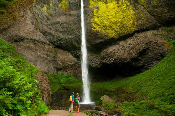 Two hikers gaze upwards at a waterfall from its base