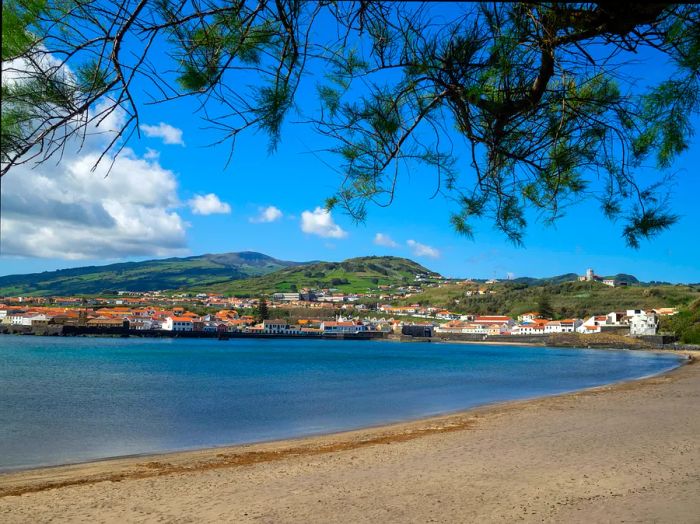 A gently curving expanse of light brown sand leads to tranquil waters, with a town visible across the bay.