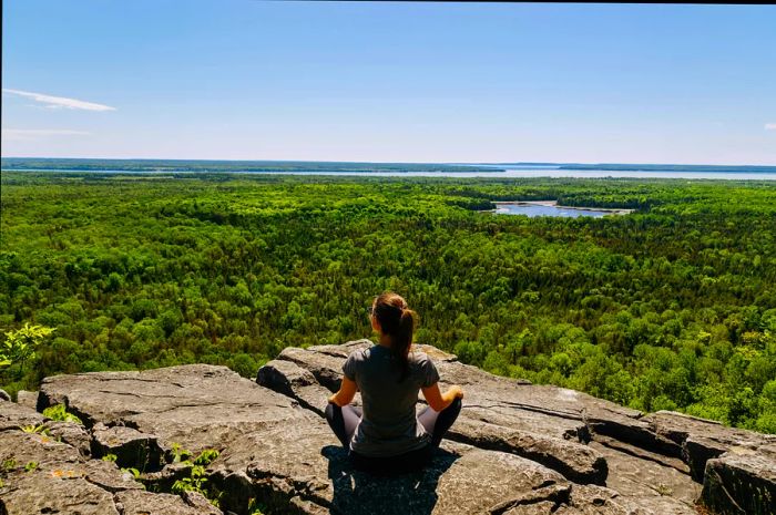 A woman perches on a rock, gazing out at the forest ahead on the Cup & Saucer Trail at Michigiwadinong, Manitoulin Island, Ontario, Canada