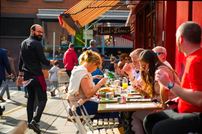 Diners enjoy meals at outdoor tables as a smiling staff member walks by