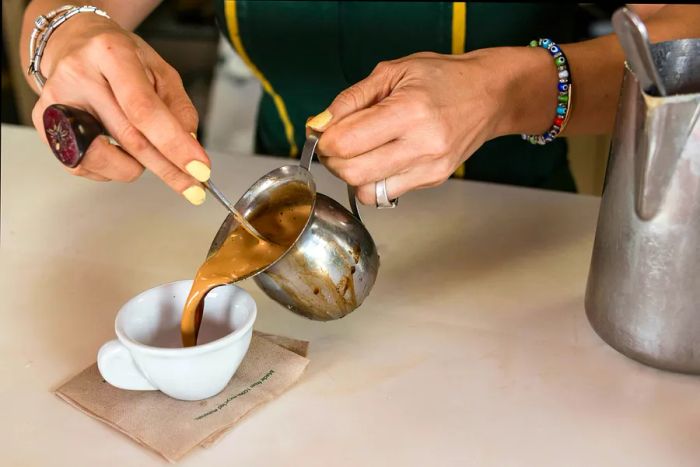 A light brown Cuban coffee is being poured from a metal jug into a small white cup.