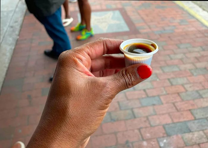 A hand holds a thimble-sized plastic cup filled with black coffee