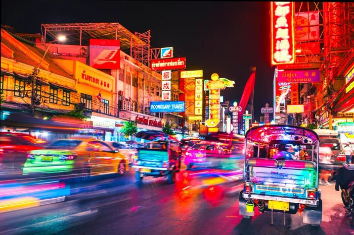 A vibrant display of neon lights as tuk-tuks and other vehicles flow through a brightly lit street at night.