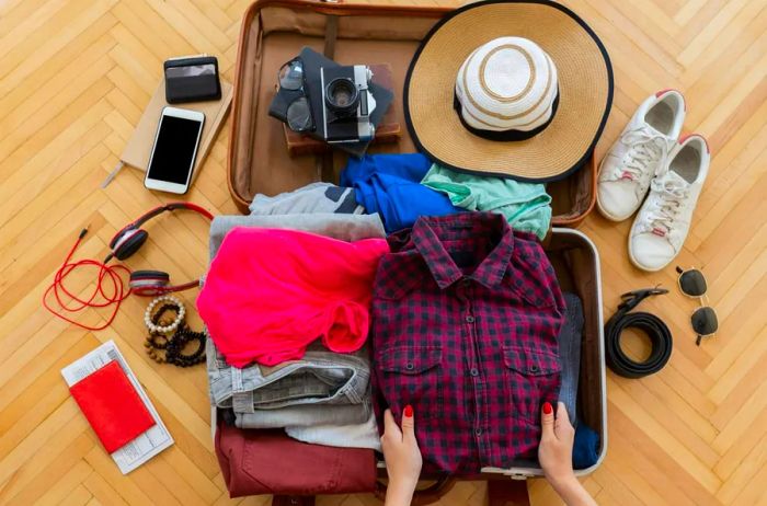 A woman’s hands prepare for a summer getaway, packing essentials like a sun hat, comfortable shoes, and various electronic devices