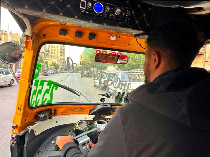 A tuk-tuk driver glances back with a small smile at his passenger in the rear-view mirror as he drives along