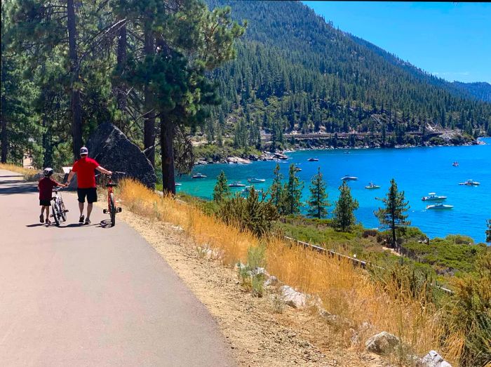 An adult and child ride their bikes along a lakeside trail.