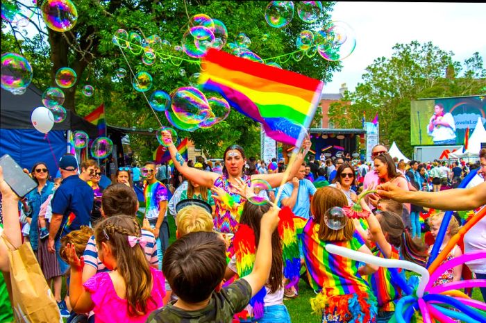 A diverse group of celebrants, including adults and children in vibrant rainbow attire, enjoy the Pride Festival in Dublin in June, waving flags and blowing bubbles.