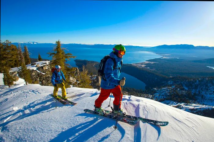 An adult and a child in full snow gear cross-country ski along a snowy ridge high above a lake