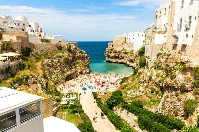 A pathway leads to a small beach bustling with visitors, nestled in a stunning cove with turquoise waters.