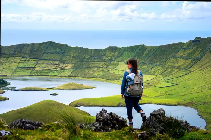 A woman stands before a green-covered crater with a lake nestled at its base.