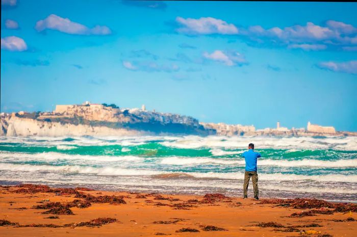 A man stands on a beach with crashing waves and captures a photo of a clifftop town in the distance.