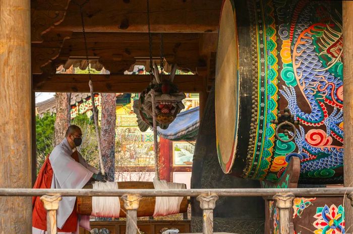 A Buddhist monk in traditional attire and a face mask stands within a temple pavilion, striking a large, ornate hanging drum with a mallet. The drum features intricate patterns, and nearby, part of a colorful dragon mural showcases the rich artistic heritage of the temple. The pavilion's architecture, with its wooden beams and decorative details, exemplifies traditional Korean style, creating an atmosphere of solemnity and cultural significance.