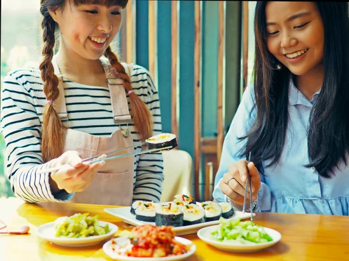 Asian women enjoying Korean seaweed rice rolls with cheese (also known as kimbap or gimbap) and kimchi, using silver chopsticks at a wooden table.