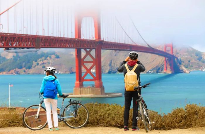 A couple on bikes stops along a trail to snap photos of a large orange-red bridge cloaked in fog.