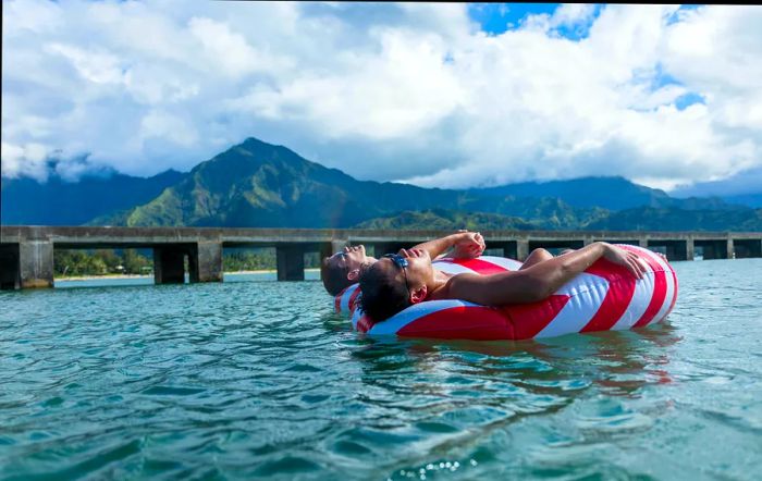 Two men share a moment, floating together in matching red-and-white striped inner tubes against a backdrop of mountains on a cloudy day.