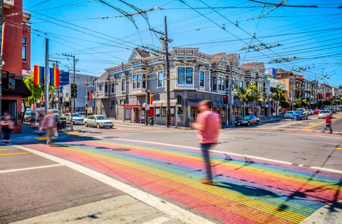 Pedestrians traverse a rainbow crosswalk in a lively neighborhood, adorned with rainbow flags from nearby buildings