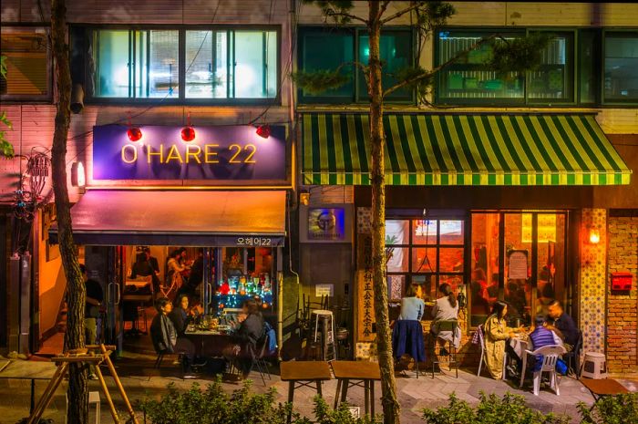 A lively street scene of patrons enjoying drinks inside and outside bars at night in Seoul.