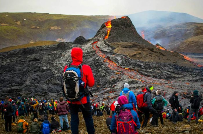 A volcano in Iceland erupts as a group of travelers watches from a safe distance.