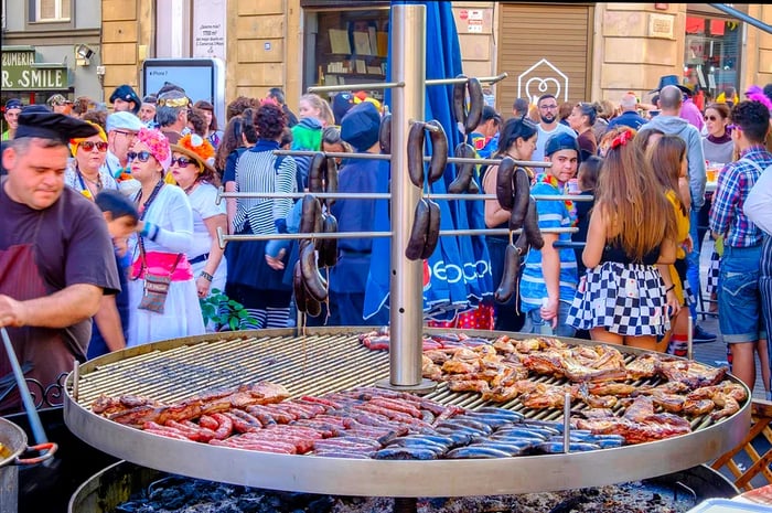 A vibrant street food market buzzing with activity, featuring a large grill cooking various meats, surrounded by a diverse crowd, including individuals in colorful costumes.