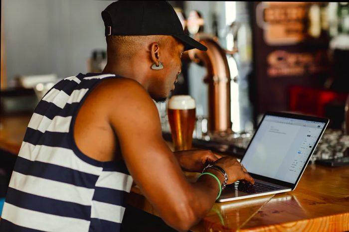 A man works on his laptop at a bar counter, enjoying a pint of beer beside him.