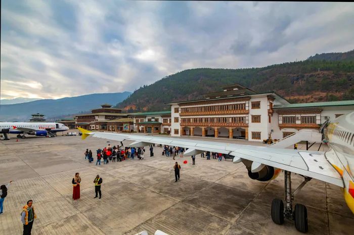 A close-up of an airplane wing with a full aircraft in the distance and people on the tarmac at Bhutan International Airport, captured during a winter daytime.