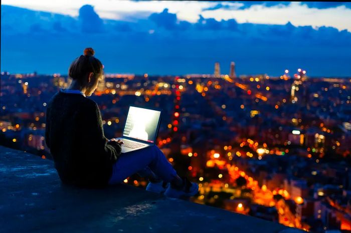 A person works on a laptop from a high vantage point at twilight, with the city lights twinkling in the background.