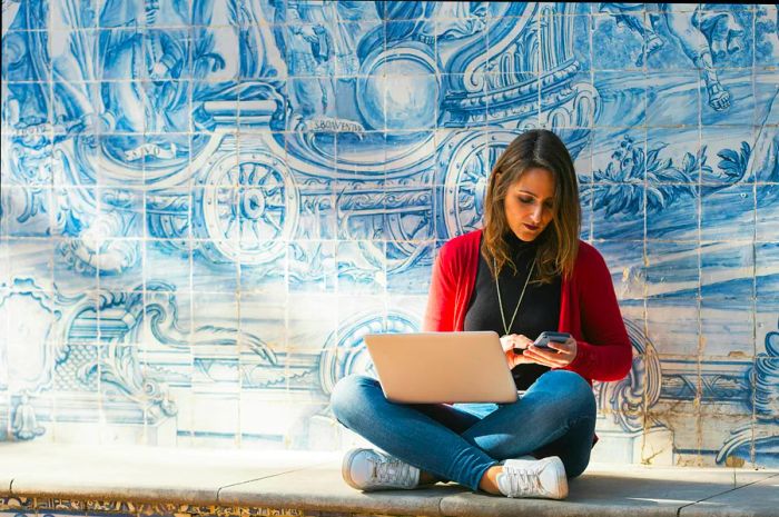 A woman is seated cross-legged, working on a laptop in front of a beautifully designed blue-tiled wall.