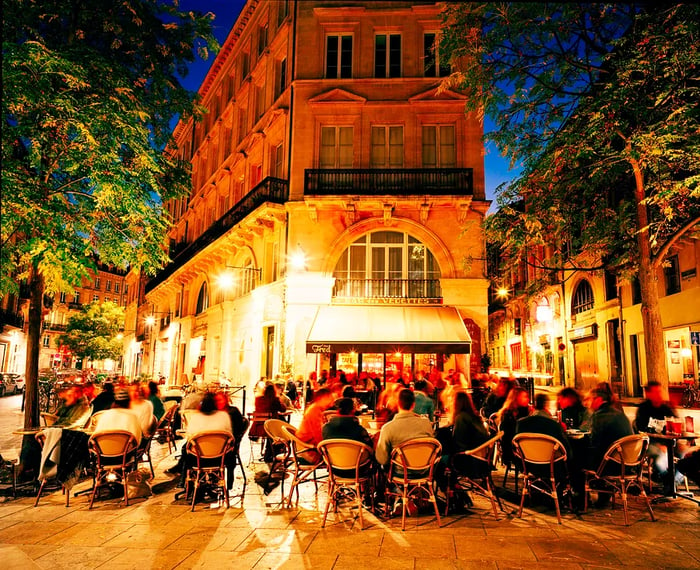 An outdoor café bustling at night, with guests gathered around tables illuminated by street lamps and a warmly lit building façade, creating a vibrant evening ambiance.