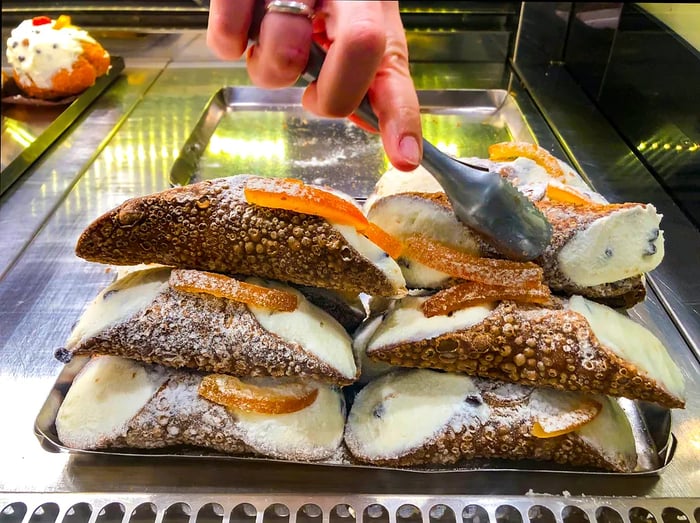 In a Sicilian pastry shop, a server presents traditional ricotta cannoli garnished with candied oranges while a pastry chef delicately arranges desserts with tongs.