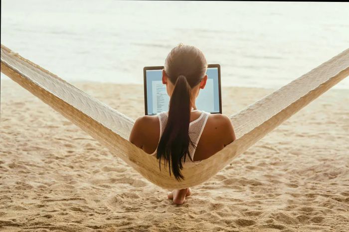 A view from behind someone lounging in a hammock with a laptop, gazing at a sandy beach.