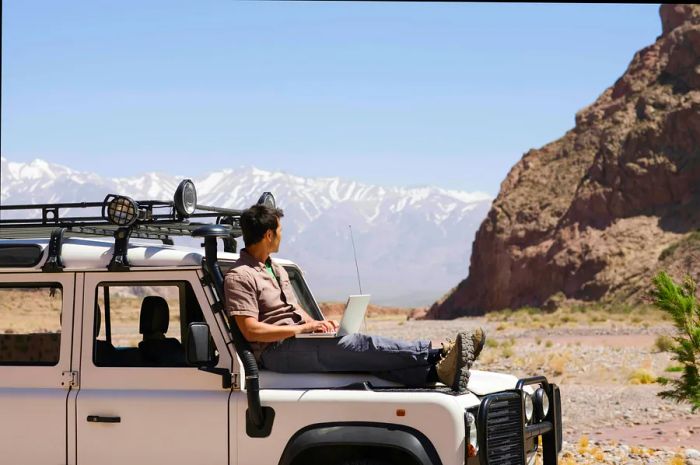 A person sits atop the hood of an off-road vehicle, working on a laptop against a backdrop of a rugged desert landscape.