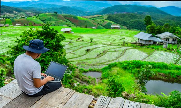 A person is working on a laptop outdoors, enjoying a view of a vibrant green landscape.