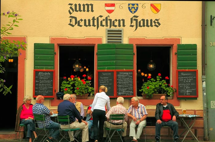 Guests enjoying their meals outside a traditional German eatery with a sign reading 'Zum Deutschen Haus', menus showcased on a blackboard, and a charming flower-adorned exterior.