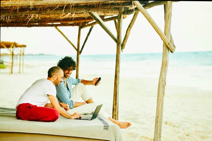 A wide shot of a smiling gay couple reviewing data on a laptop while lounging at a beach cabana in a tropical resort.