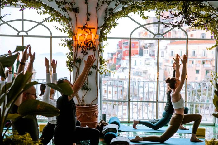 A group of individuals practicing yoga in a spacious room with large windows showcasing a historic city view, bathed in natural light and adorned with decorative plants in the foreground.