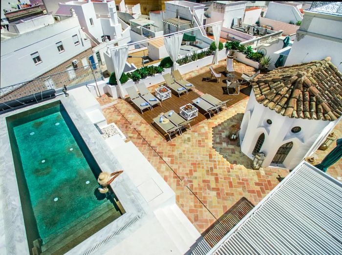 An aerial perspective of a rooftop terrace featuring a swimming pool, sun loungers, and umbrellas, with the charming town and its buildings visible in the background.