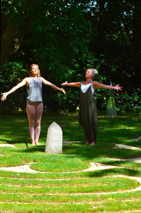 Two women in a garden with arms outstretched towards each other, a large clear crystal resting on the ground between them, surrounded by lush green trees.