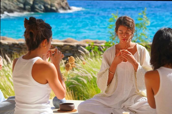 A woman sits cross-legged in meditation on a stone terrace by the sea, facing another person, both enveloped by a natural landscape and a clear blue sky.