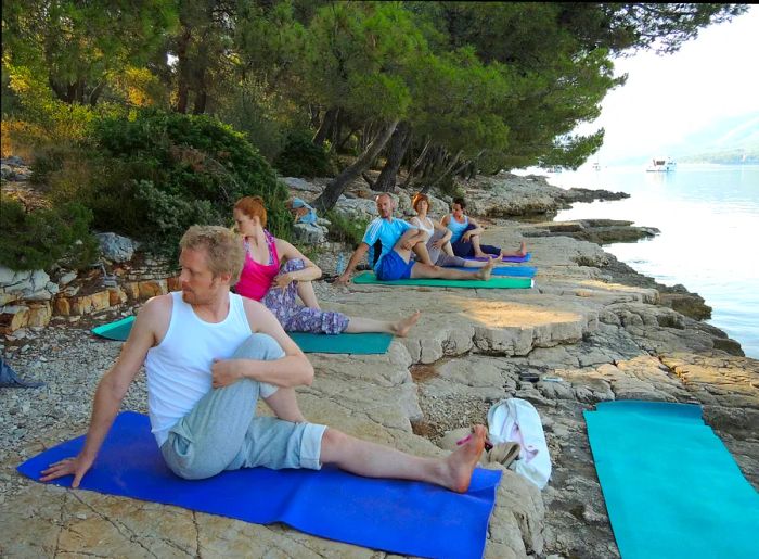 A group of people participating in a yoga class on a rocky beach by the sea, with pine trees in the background and a distant boat visible.