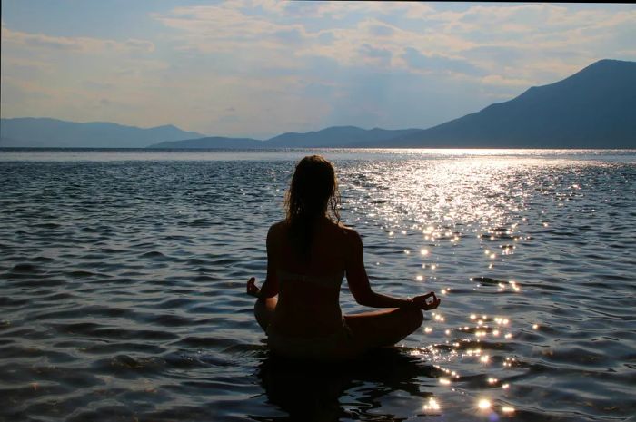 A silhouette of an individual meditating in the lotus position by the sea at sunset, with mountains in the background and the water shimmering in the light.