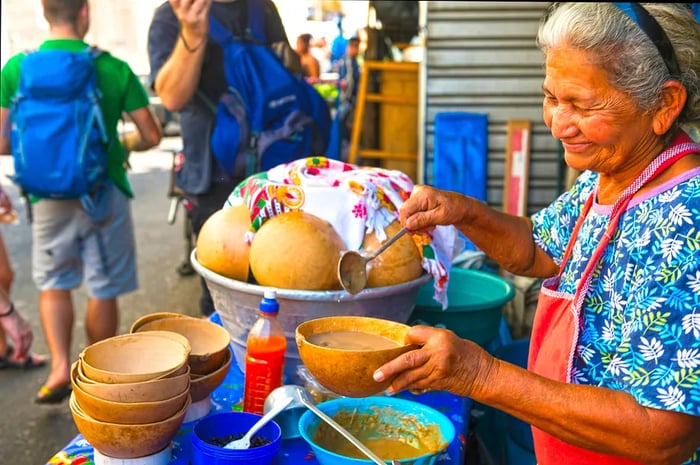 An elderly woman cheerfully pouring soup into a pumpkin bowl at her street food stall, with people in the background.