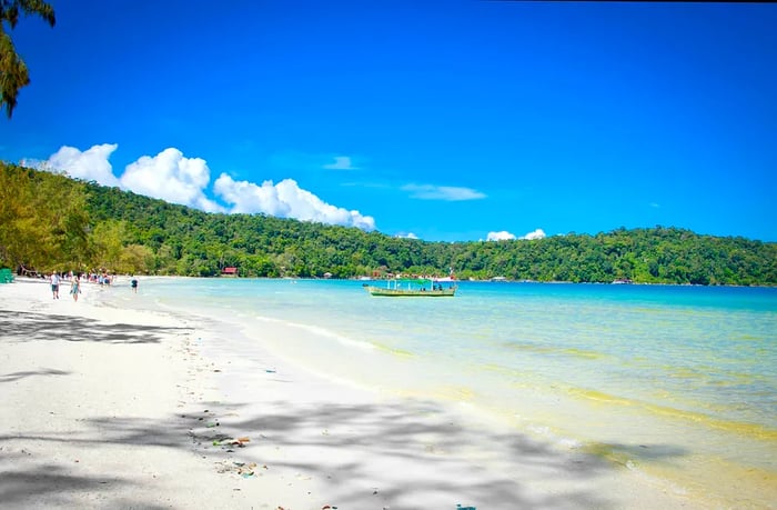 A broad white-sand beach dotted with visitors and a small boat, set against a backdrop of turquoise waters and a low green mountain range in the distance