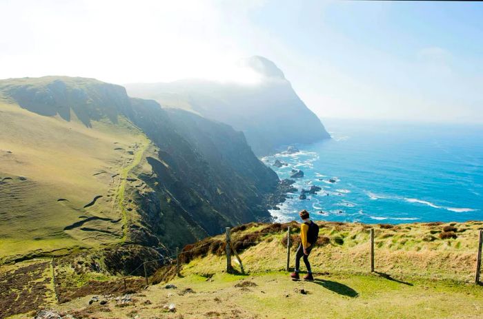 A hiker stands atop a grassy cliff, gazing at the ocean, with a mountain partially shrouded in mist and a clear blue sky overhead.