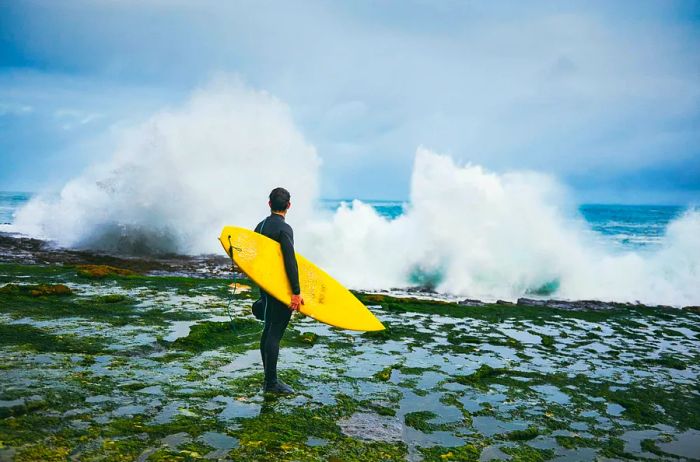 A surfer in a full wetsuit grips a yellow surfboard, gazing at the massive Atlantic waves crashing against the Irish coast.