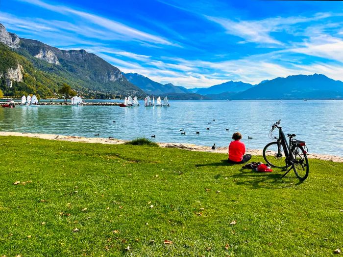 A lone cyclist relaxes beside their bike by the lakeside, enjoying the view of the shimmering waters and distant sailboats.