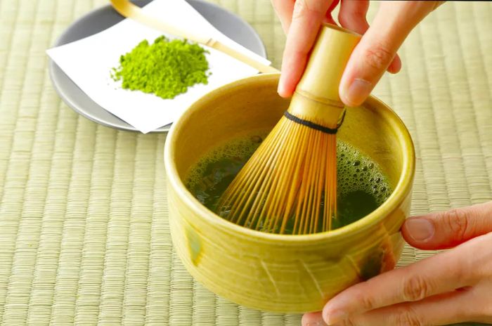 A person prepares green tea in a small bowl using a bamboo whisk, with powdered matcha resting nearby on a plate.