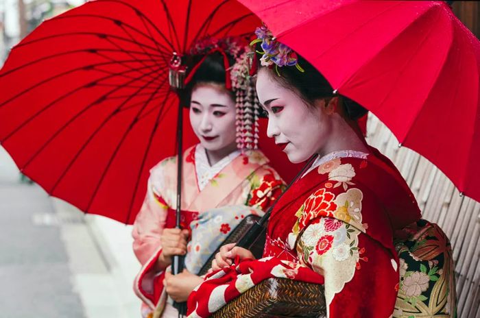 Two geisha dressed in traditional garb stand under red umbrellas on a Gion street.