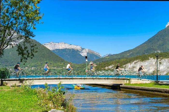 A group of six cyclists ride across a bridge next to a lake surrounded by mountains