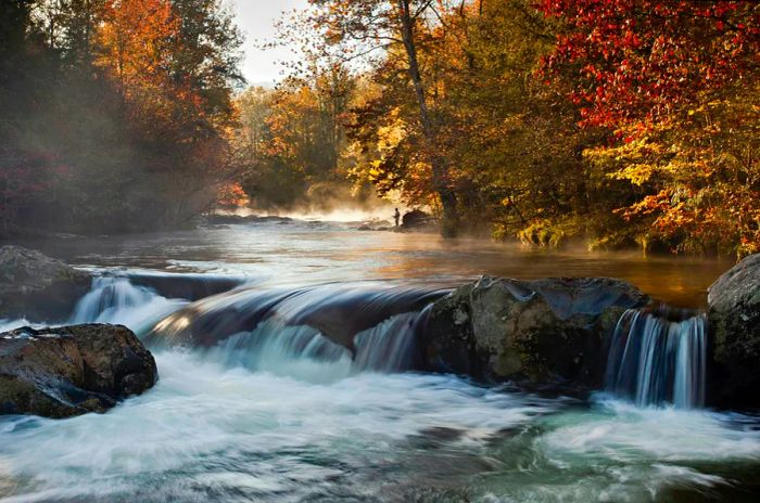 A fisherman stands amidst an autumn landscape, with mist hovering over the river and water cascading over rocks in the foreground.